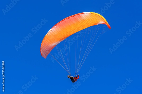 Fototapeta A single paraglider with a vibrant orange and red canopy is seen against a deep, clear blue sky during a sunny day flight.
