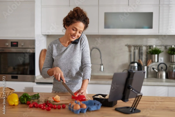 Fototapeta In a bright kitchen, a woman is chopping vegetables while engaged in a phone conversation. Fresh produce surrounds her, adding color to the scene.