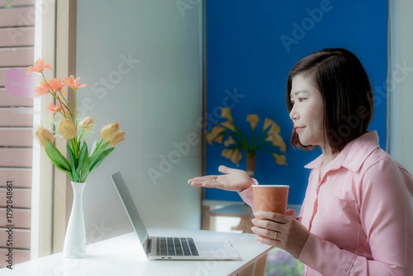 Fototapeta Businesswoman smiling while using laptop and talking on mobile phone at desk in office and at home or coffee shop.