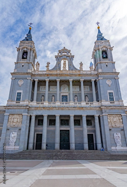 Obraz The Almudena Cathedral in Madrid, showcasing its neo-Romanesque exterior, with twin bell towers, a central dome, and a large stone staircase under a dramatic blue and white sky.