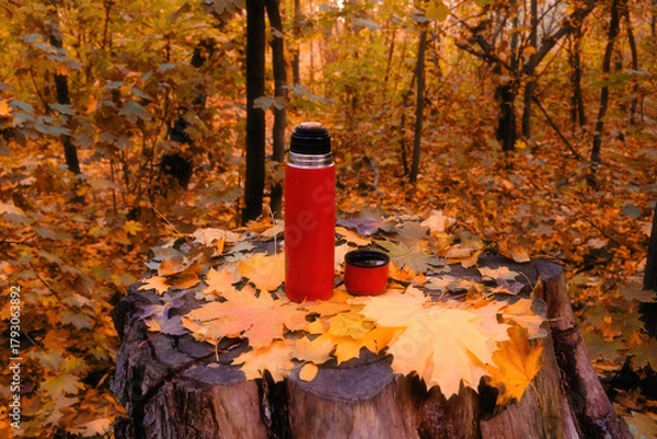 Obraz Autumn warmth, a red thermos with hot tea and a mug on a tree stump, among bright yellow leaves in a sunny forest. The perfect moment for coziness and outdoor recreation.