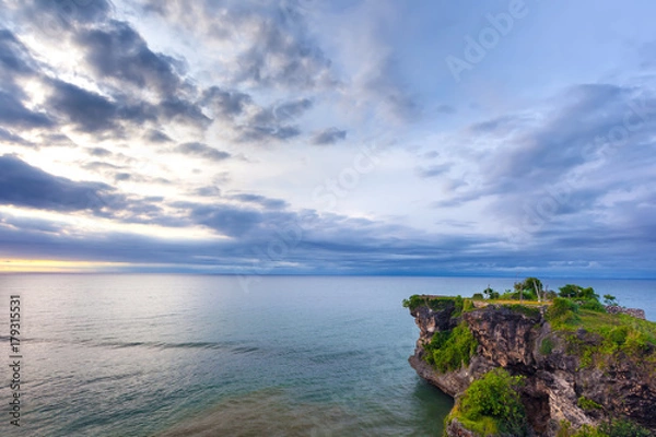 Fototapeta Volcanic green rock with huge stones, on the sea shore, against the backdrop of beach sunset. Beautiful blue clouds on the horizon. Balangan beach view, Jimbaran, South Kuta, Bali, Indonesia.