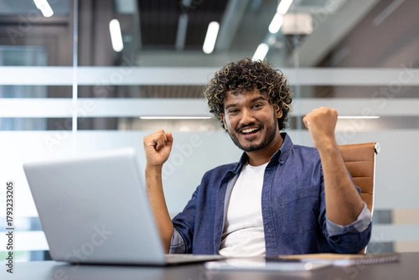 Fototapeta Portrait of a happy young Indian man sitting in the office at a table with a laptop, raising his hands up and rejoicing