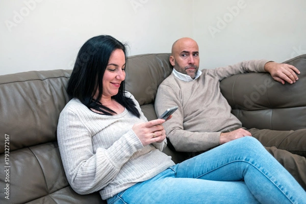 Obraz A woman smiles while using her smartphone on a sofa, as a man beside her looks on with a serious, uneasy expression. The scene captures tension and mixed emotions in a domestic environment.