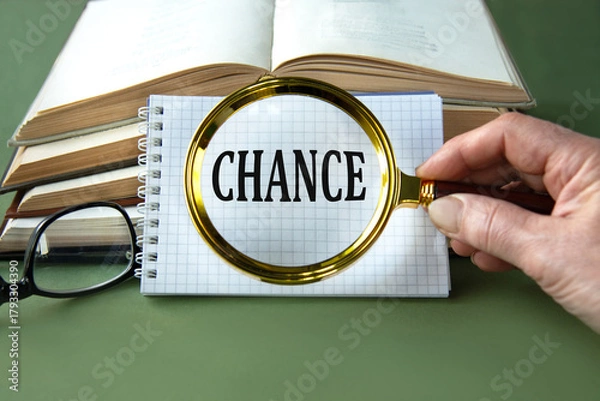 Fototapeta A woman's hand holds a magnifying glass over the word CHANCE on a green background with books