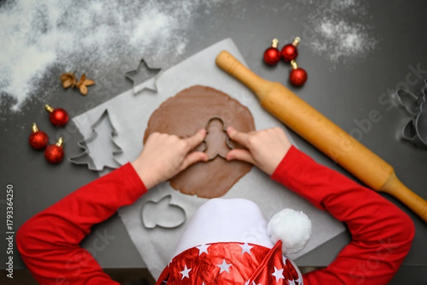 Fototapeta Top view of child making gingerbread cookies with Christmas mold on table, surrounded by festive decorations. Perfect for winter marketing, cozy home baking concepts, and joyful holiday designs.