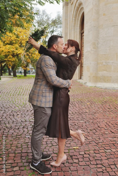 Obraz Romantic stylish couple sharing a deep kiss and holding a bouquet of white roses on a brick cobblestone street in autumn