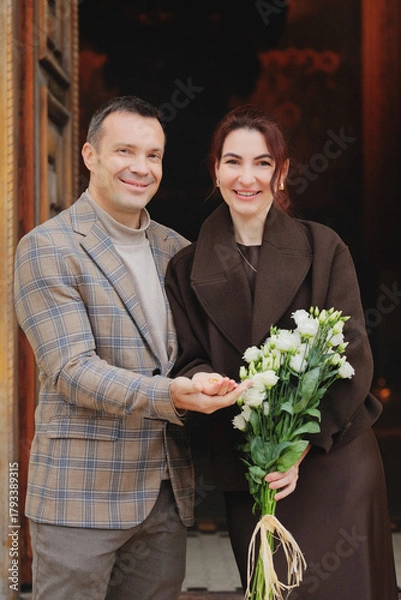 Obraz Happy elegant couple smiling at the camera, holding hands and a bouquet of white flowers in front of a dark, historic entrance