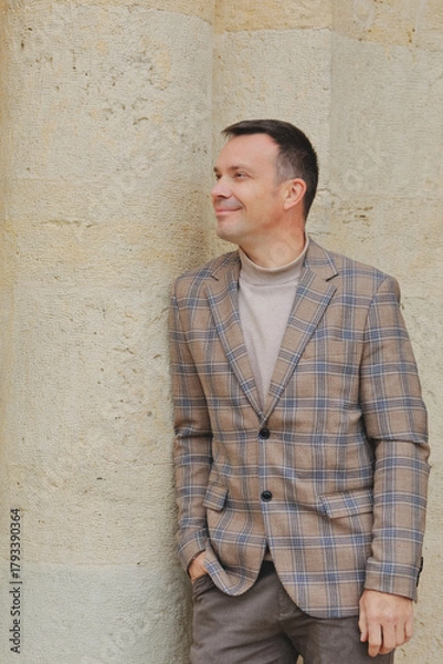 Fototapeta Happy, stylish middle-aged man in a plaid blazer and turtleneck, leaning casually against an old limestone wall and looking away