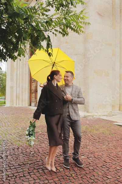 Fototapeta Happy elegant couple smiling at each other under a bright yellow umbrella, holding white roses on a red cobblestone street in autumn