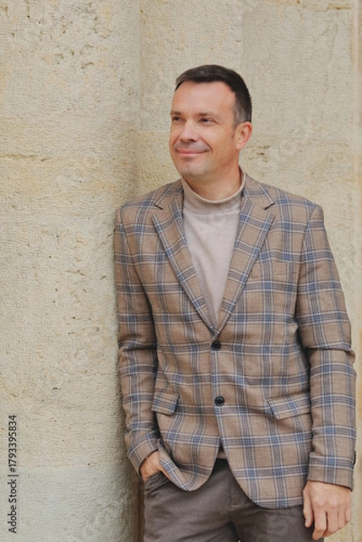 Fototapeta Happy, stylish middle-aged man in a plaid blazer and turtleneck, leaning casually against an old limestone wall and looking away