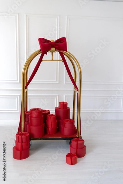 Obraz Elegant hotel luggage cart decorated with a big red bow and filled with red gift boxes. Festive holiday or luxury celebration concept in bright, minimal interior