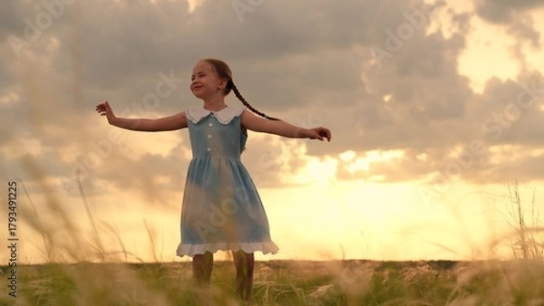 Obraz Happy child girl plays spinning in park on grass. Child girl in dress dances spinning with her arms out to sides. Child dream to dance. Little girl smiles in front of sky. Children's dreams in outdoor