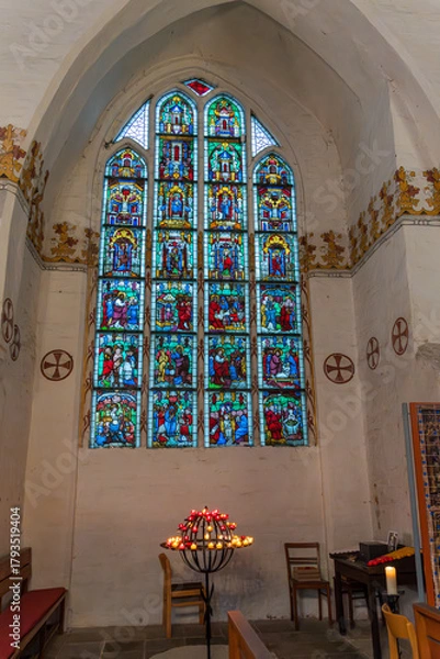 Fototapeta Colorful stained-glass windows depicting biblical scenes adorn a chapel, with candlesticks and simple wooden pews in front.