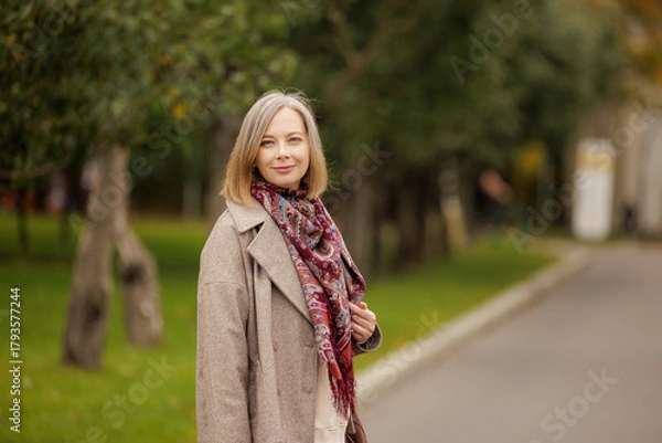 Fototapeta Smiling woman in a cozy scarf enjoys a peaceful stroll through a serene park on a beautiful autumn day