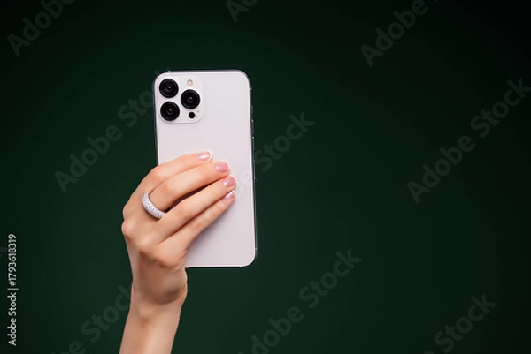 Fototapeta Woman hand holding a modern smartphone showcasing style, technology, and luxury against a rich green background