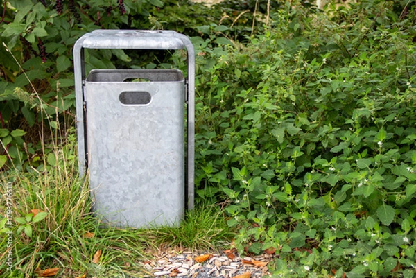 Fototapeta Metal trash bin stands amidst lush green foliage, surrounded by grass and small stones, showcasing an eco-friendly waste disposal solution in a natural environment