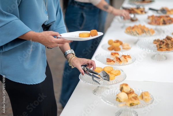 Fototapeta Guests select delicious food from an elegant buffet table during a private event showcasing a variety of appetizers and treats presented artfully on clear glass platters ideal for social gatherings