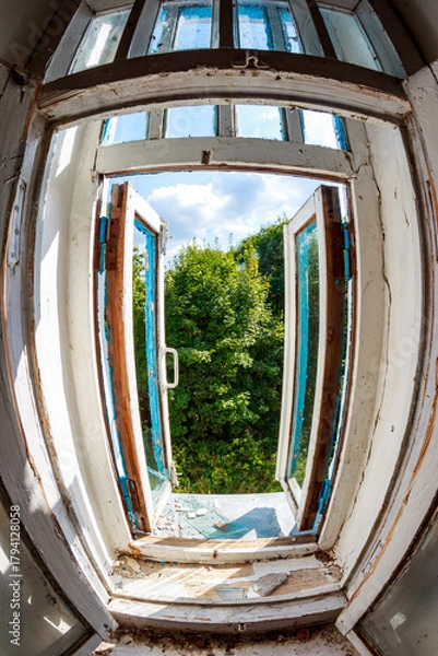 Fototapeta Dilapidated wooden window frame inside abandoned building, wide-angle view. Open panes reveal vibrant summer greenery and sky. Broken glass, peeling paint, urban decay