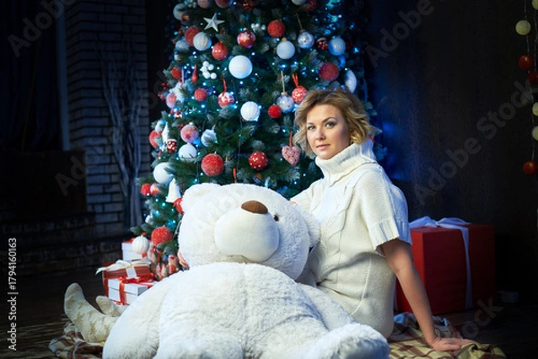 Obraz A woman in a white sweater is sitting on the floor with a white teddy bear near a Christmas tree.