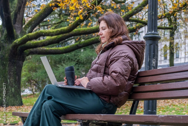 Fototapeta Midlife woman engaged in video call meeting while working remotely on laptop in a park, surrounded by autumn foliage and natural scenery, showcasing modern work-life balance