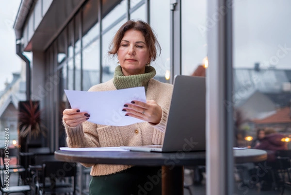 Fototapeta Middle-aged woman engaged in remote work at a food court, reviewing documents while seated at a table with a laptop and a cozy atmosphere, showcasing modern work-life balance
