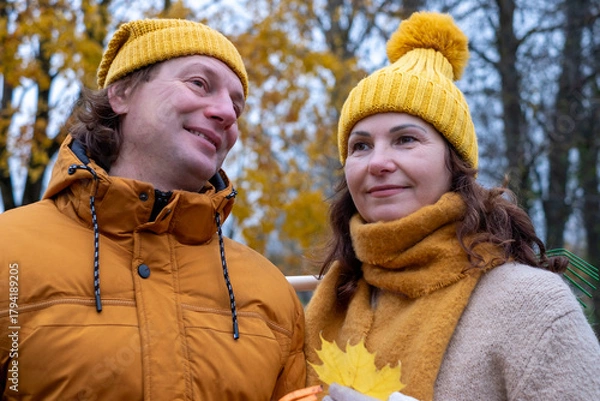 Fototapeta Middle-aged couple in cozy autumn attire smiles while holding colorful leaves, surrounded by vibrant fall foliage, enjoying a peaceful moment in nature during the autumn season