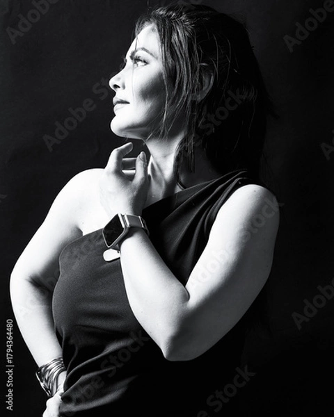 Fototapeta Black and white fine-art portrait of a woman in a one-shoulder dress, wearing wrist accessories and looking upward under studio lighting. Perfect for concepts of confidence, introspection, fashion