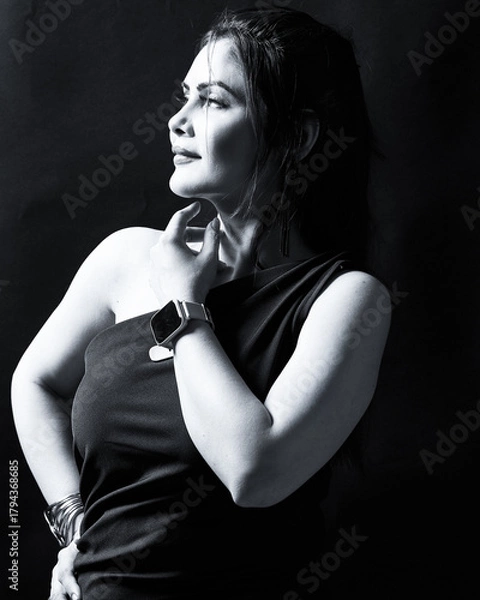 Obraz High-contrast monochrome portrait of an elegant woman in a one-shoulder dress and wrist accessories, looking upward with a confident expression. Ideal for editorial, fashion, and empowerment themes.
