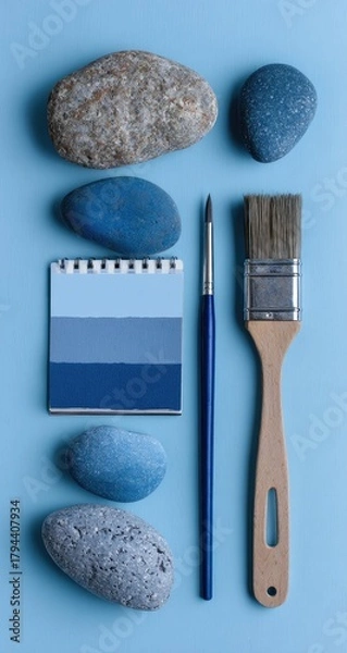Fototapeta Flatlay of painted stones, paintbrushes, and a color palette on a light blue background.  The arrangement is vertical and symmetrical