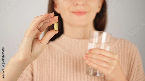 Obraz Macro close-up of an unrecognizable smiling woman holding vitamin capsule and glass of water. Advertising concept for vitamins and nutritional supplements, health care.