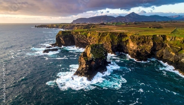 Fototapeta Rocky coastline with cliffs and mountain backdrop under cloudy sky, ocean waves crashing