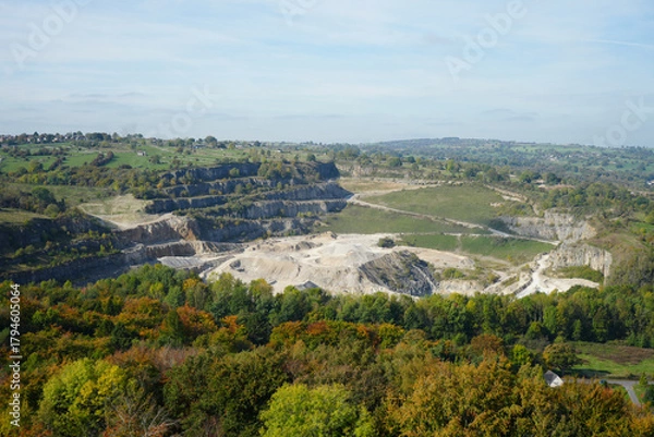 Fototapeta Panoramic View Of An Active Quarry At Black Rocks, Cromford, Derbyshire, Showing Terraced Rock Formations, Autumn Trees, And Distant Countryside Under A Clear Sky.