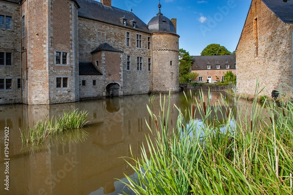 Fototapeta A scenic view of Château de Lavaux-Sainte-Anne in Belgium with its medieval walls rising above a peaceful moat, surrounded by greenery and bathed in warm sunlight under a clear blue sky.