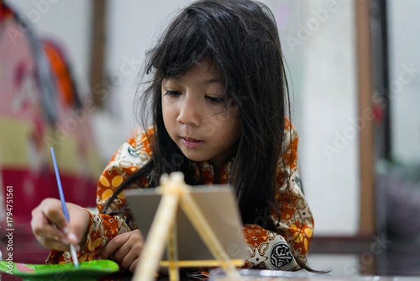 Fototapeta A young girl focuses while painting on a small canvas using a brush. She wears a colorful batik outfit as she works creatively at home, showcasing a genuine moment of childhood art and imagination.