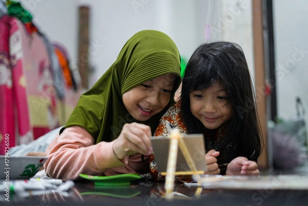 Obraz Two young girls painting together on a small easel while lying on the floor at home. They look focused and happy as they share a creative activity using brushes and bright colors.