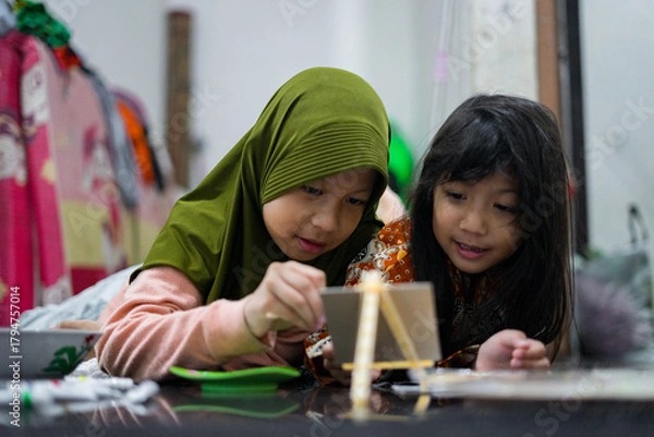 Obraz Two young girls painting together on a small easel while lying on the floor at home. They look focused and happy as they share a creative activity using brushes and bright colors.
