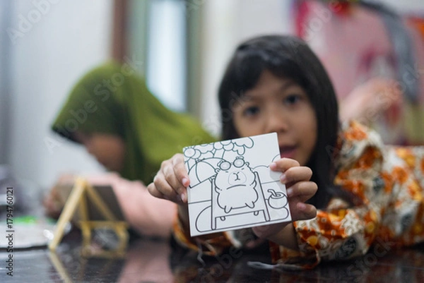Obraz A young girl in batik clothes shows a small drawing board with a cute cartoon picture while lying on the floor. Another child in a green hijab is painting in the background.
