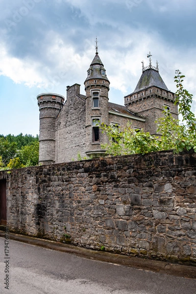 Fototapeta Spontin Castle Château De Spontin rises beyond its tall stone wall, showcasing pointed towers and ornate turrets under a dramatic cloudy sky in the Belgian countryside, rich with medieval heritage and