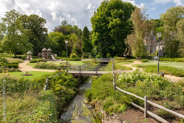 Fototapeta Lush garden landscape near Spontin Castle in Belgium featuring wooden bridges, flowering bushes, tall trees, and a playground under a partly cloudy sky creates a peaceful and charming countryside retr