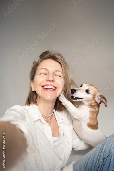 Fototapeta Happy friends selfie. blond woman and small dog Jack Russell terrier. woman smiling looking at camera and touching her face.  Dog owner and pet having fun in studio grey background. empty copy space
