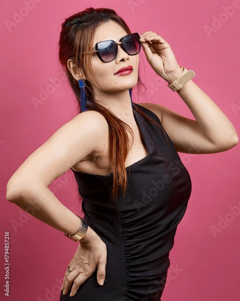 Fototapeta Studio fashion portrait of a confident woman in a black dress wearing sunglasses against a pink background. Ideal for concepts of fashion, style, empowerment, and summer elegance.
