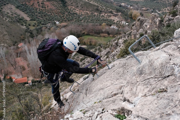 Fototapeta A climber ascends a steep rocky wall on a via ferrata in Castillo de Locub n, Ja n, Spain, using safety cables and metal steps surrounded by olive groves.