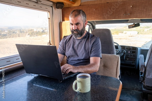 Fototapeta A middle-aged bearded man types on his laptop inside a camper van, with natural light entering through the window and a mug of coffee on the table in front of him.