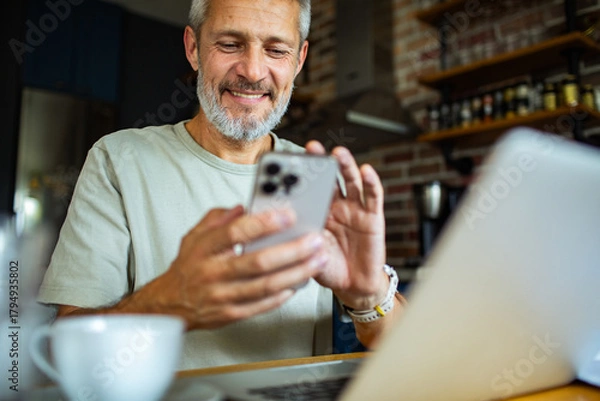 Fototapeta Mature man smiling and using smartphone in home kitchen