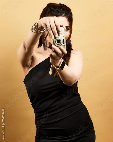 Obraz Studio portrait of a stylish woman in a black dress holding a camera toward the viewer against a golden background. Symbolizes creativity, empowerment, and confidence in modern fashion.