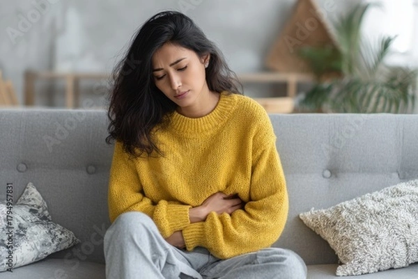 Fototapeta Distressed Indian woman clutching her abdomen experiencing cramps while seated on a couch showing discomfort and unhappiness with extra space for text