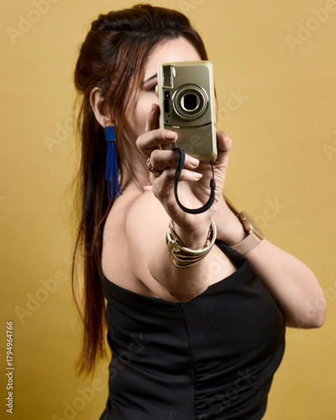 Obraz Studio portrait of a stylish woman holding a camera toward the viewer, set against a golden backdrop. Represents empowerment, creativity, and self-confidence in a modern fashion style.