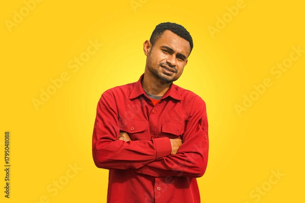 Fototapeta A black young man stands with folded arms against a yellow background, expressing a mix of confidence and curiosity. Studio portrait with bold colors, suitable for advertising and lifestyle themes.