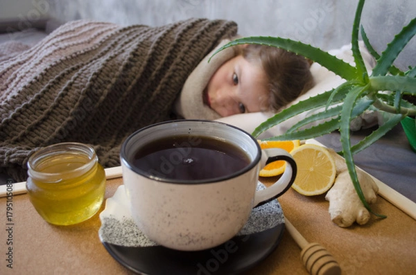 Fototapeta A little girl lies in bed drinking tea with lemon. Concepts of illness, cold, and acute respiratory viral infection.